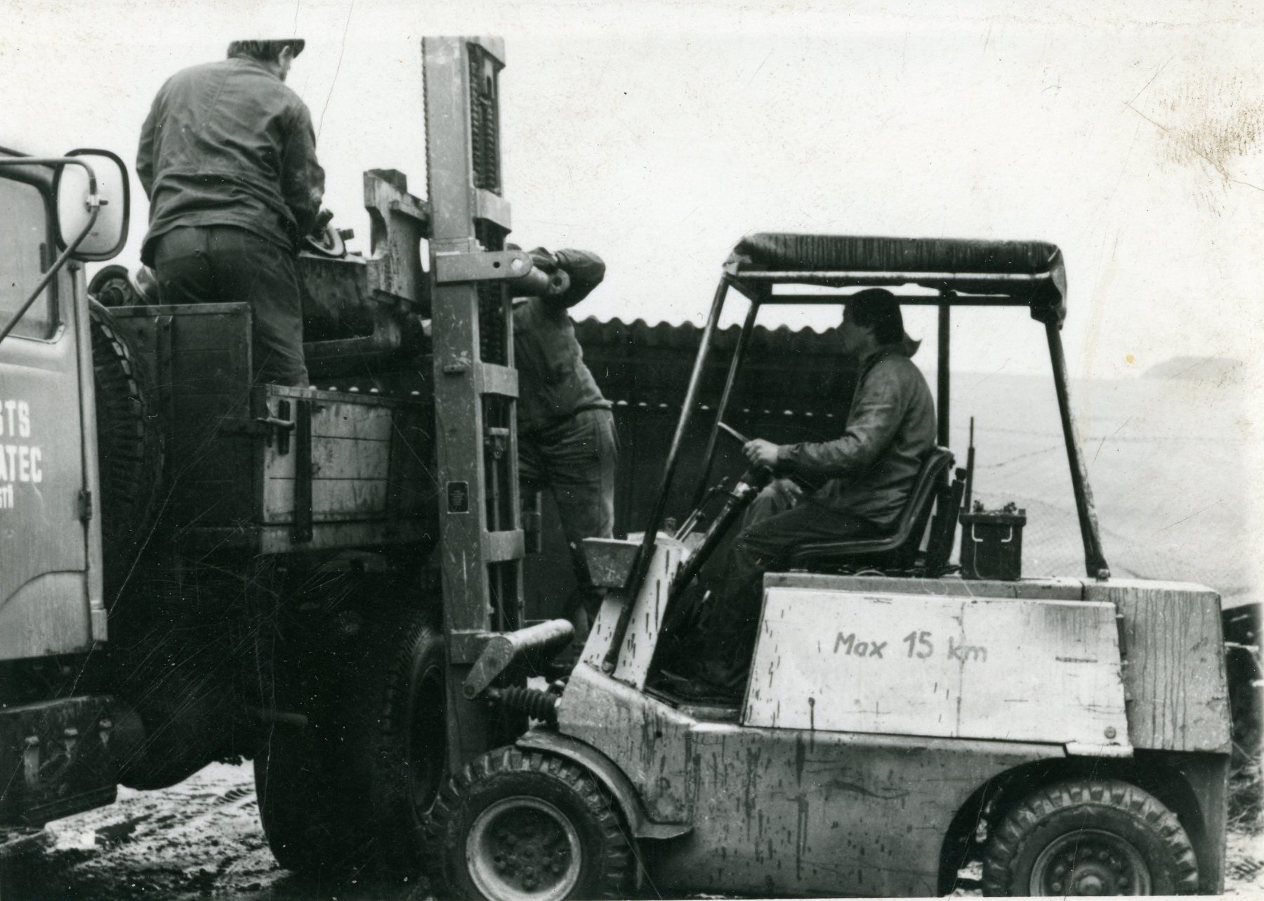 Dobová černobílá fotografie z roku 1975 — vysokozdvižný vozík nakládá techniku na nákladní Praga V3S STS Žatec. Foceno v místě, kde dnes stojí kovárna Otevřené dílny Vroutek.