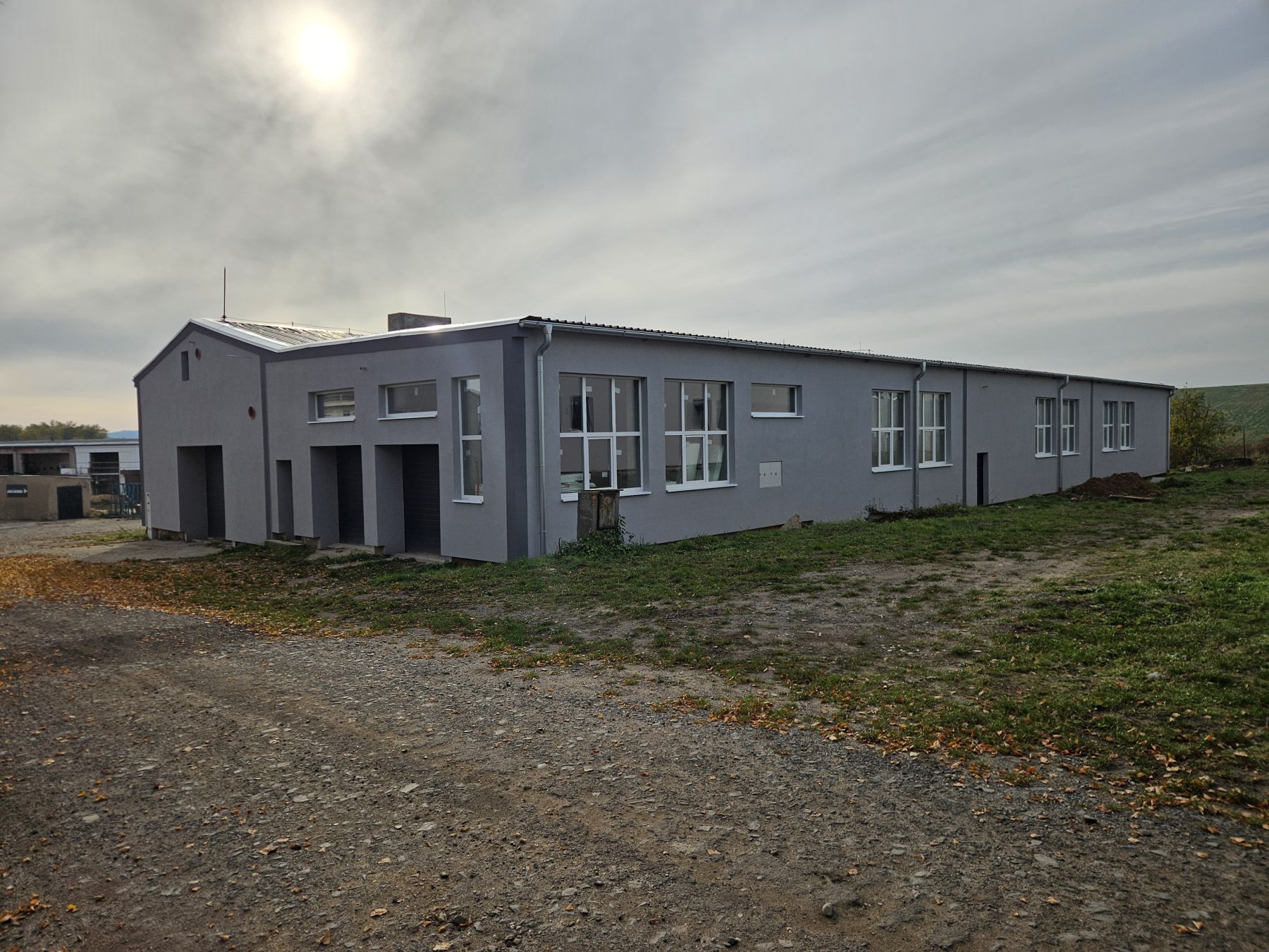 Pohled zvenku na zrekonstruovanou budovu všeobecné dílny Otevřené dílny Vroutek — dlouhá jednopodlažní hala se šedou fasádou, řadou velkých oken a sekčními vraty na čelní straně, štěrkový příjezd v popředí.